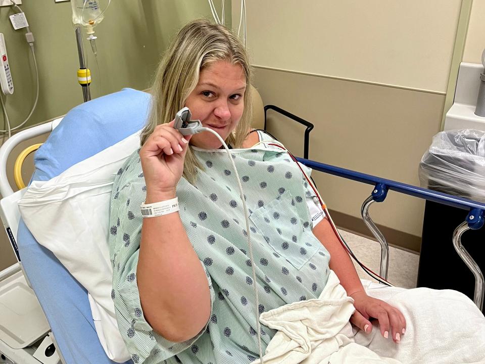 Terri Peters sitting in a hospital bed, wearing a hospital gown and wearing a monitor on her right index finger. She sits angled towards the viewer's right and poses with her right hand raised. She presses her lips together, looking slightly nervous but excited.