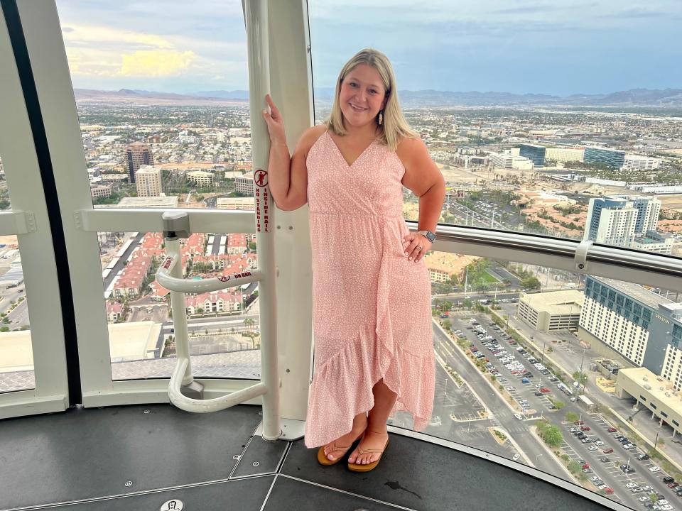 Terri Peters stands against a giant glass window with a cityscape behind her. Terri has chin-length blonde hair with dark roots, brown eyes, and wears a pink strappy dress with ruffles along the edge and tan flip-flops.