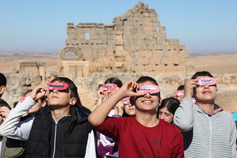 huddle of people look up toward the sun while holding pink eclipse glasses up to their eyes in front of a wall of sand colored bricks in desert landscape