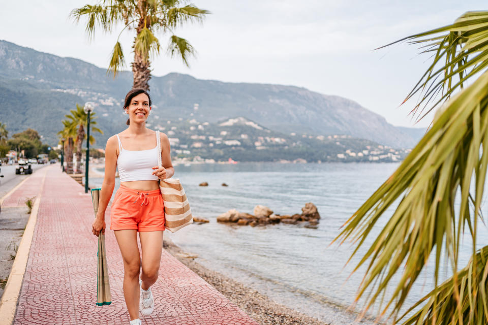 A woman wearing a white tank top and orange shorts is walking along a seaside promenade, carrying a beach bag. Mountains and palm trees are in the background