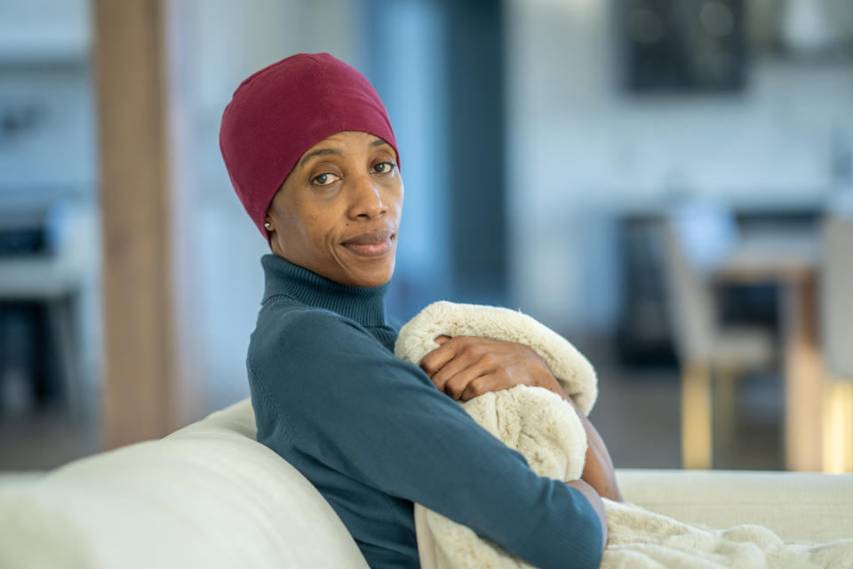 Woman in a beanie sitting on a couch, embracing a blanket, and looking pensive