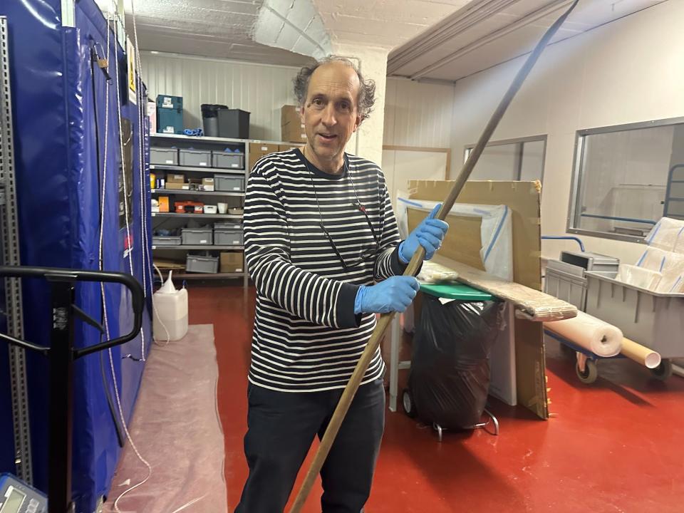 man in black and white striped shirt wearing blue medical gloves holds a long arced wooden bow in a basement room full of storage bins and packing supplies