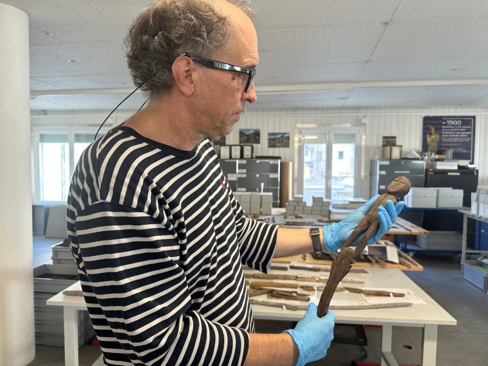 archaeologist wearing glasses black and white striped shirt and blue medical gloves holds a wooden armless human figure