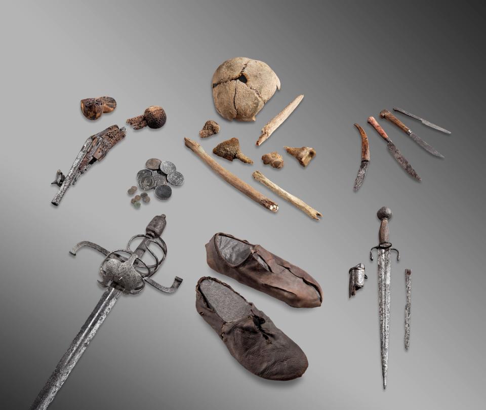 A skull cap, small bones, rusty knives, a dagger, a sword, coins, a broken pistol, and worn leather shoes spread out on a gray background.