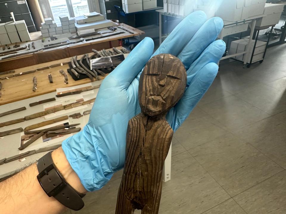 wooden human figure with no arms and a flat almost frowning expression with its head about the size of the palm of a blue medical gloved hand holding its head