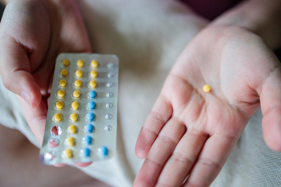 A hand holding a blister pack of pills and the other hand holding a single yellow pill between the thumb and index finger