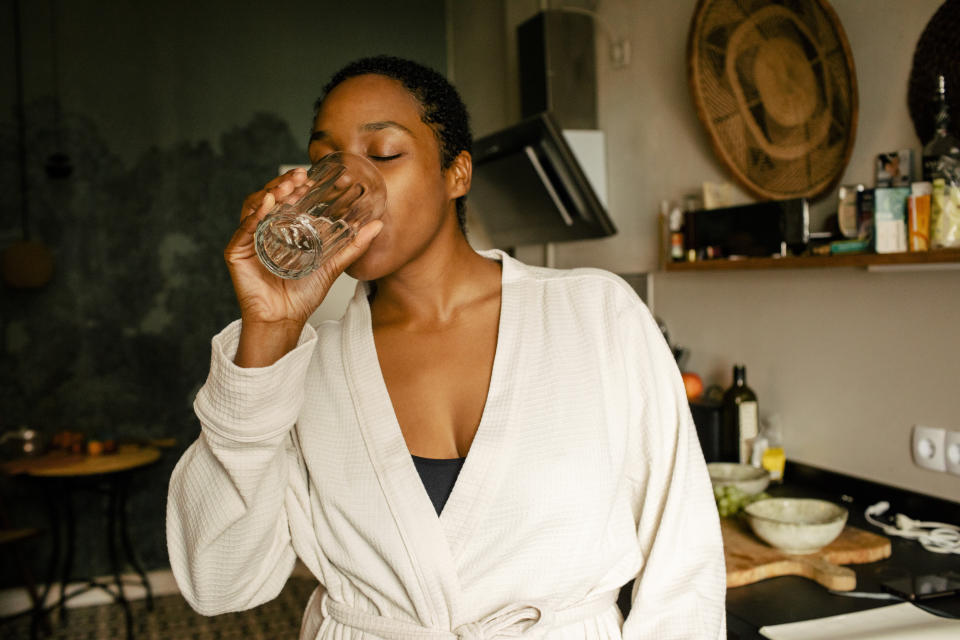 Person wearing a bathrobe, standing in a kitchen and drinking water from a glass