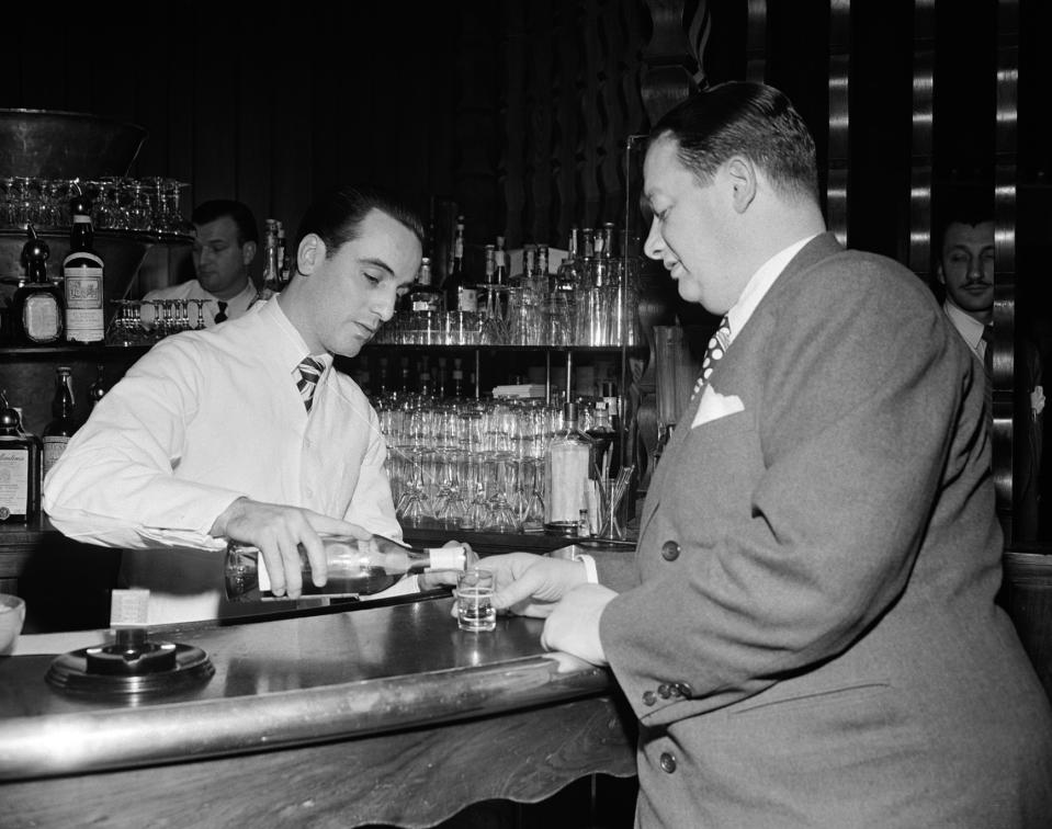 Bartender pours a drink for a customer at a bar. Both men are dressed in formal attire. Several bottles are displayed on shelves behind the bar