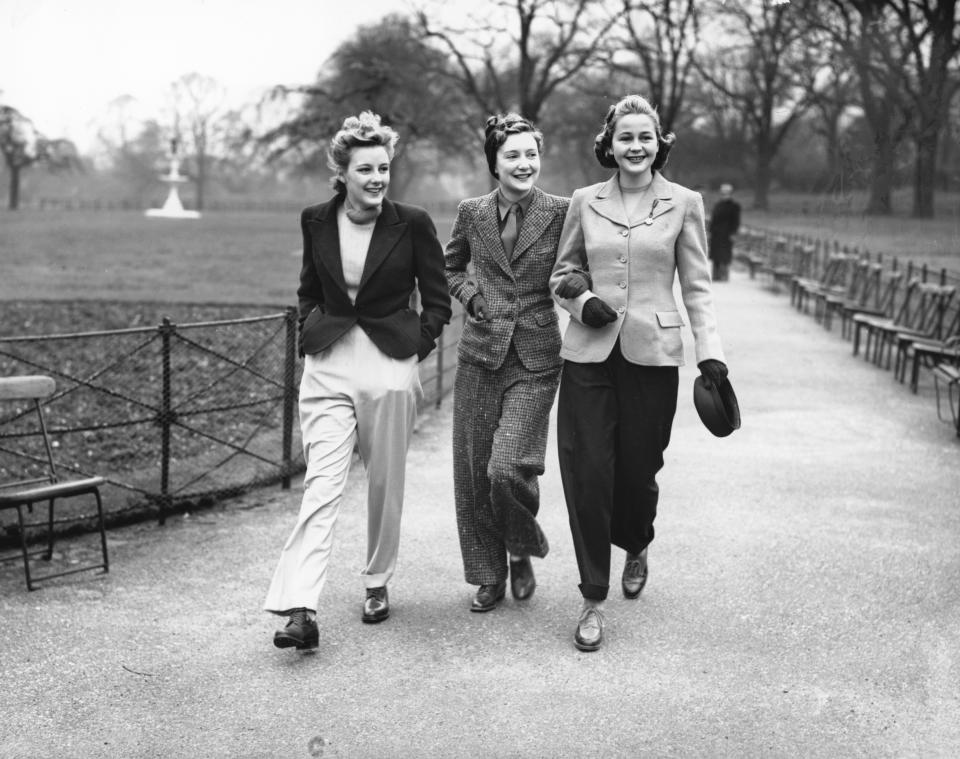 Three women walk together in a park, dressed in 1940s-style clothing with tailored jackets and loose trousers