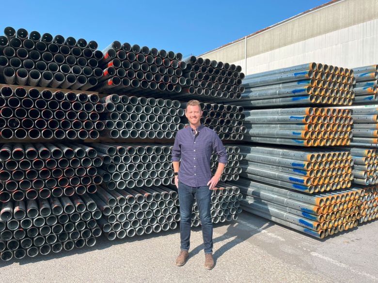 a man in jeans and collared shirt stands in front of a tall stack of thin steel pipes