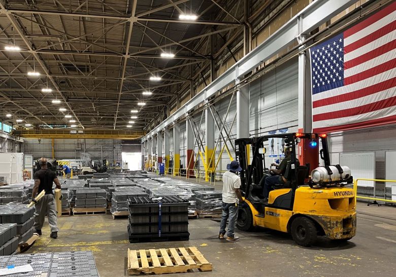 A factory floor with equipment and workers and a big American flag suspended from the wall.