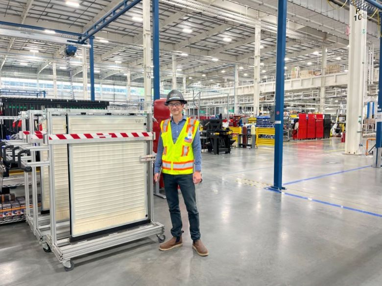 A man in a hardhat and bright, yellow vest stands in front of a battery cell.