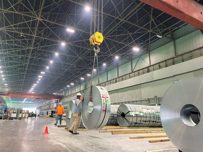 A man in a factory helps guide a large, suspended coil.