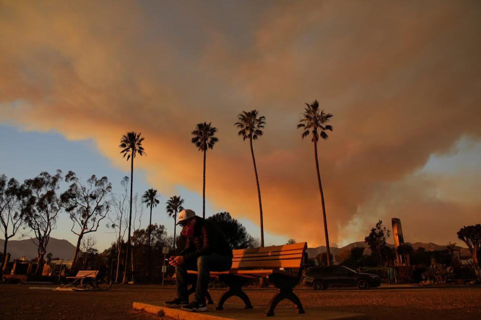 A person sits on a bench as a plume of smoke rises from the Palisades Fire on Friday, Jan. 10, 2025, in the Pacific Palisades section of Los Angeles.