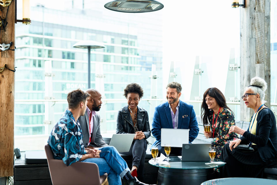 A diverse group of six colleagues are engaged in a lively discussion on a rooftop patio, laptops open, suggesting a collaborative work meeting