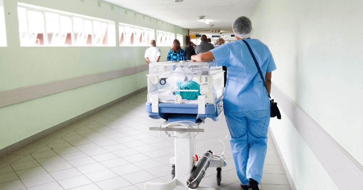 A nurse walking with an infant in an incubator down a hospital hallway.