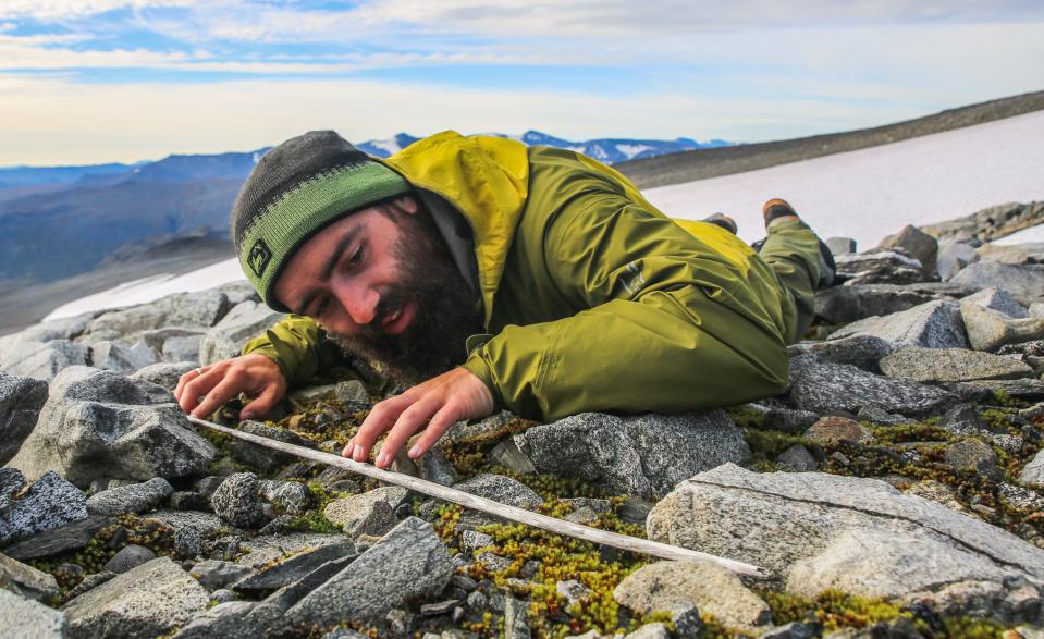 man wearing green jacket and mountaineering hat laying on his stomach in a field of rocks admiring and lightly touching his fingertips to a long thin wooden arrow shaft lain across the rocks against a background of snow and mountains