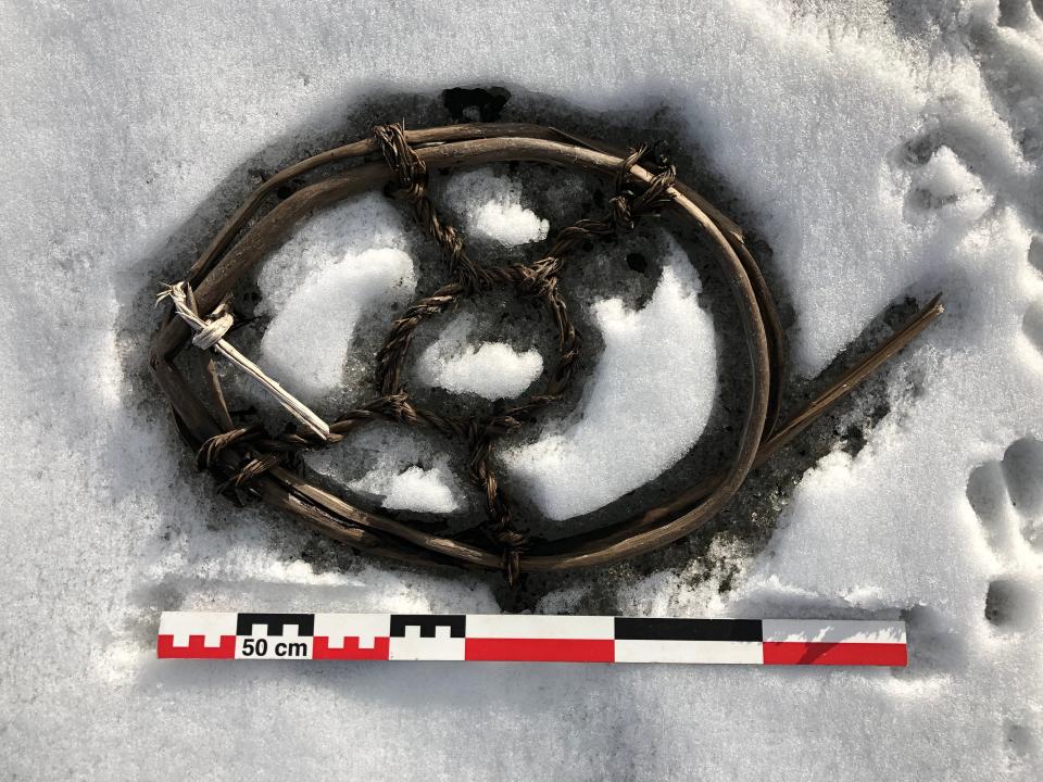 wood sticks bent in an oval and bound with spokes of ropy material sits in the snow next to a ruler showing it's about 500 cm across