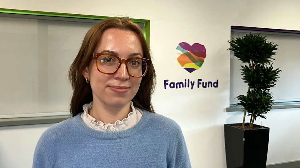 A young woman with brown hair and glasses, a light blue jumper smiling at the camera in an office environment. In the background is a Family Fund logo and a potted plant. 