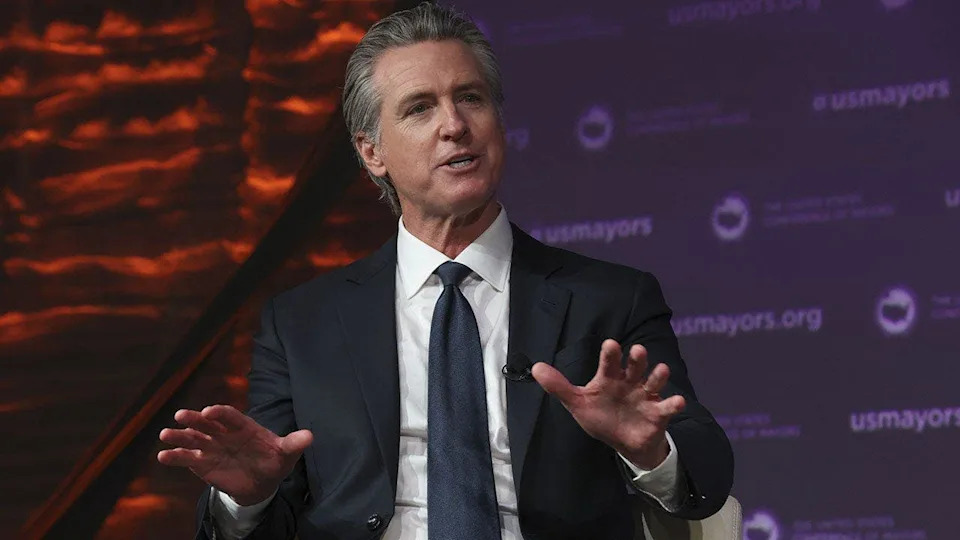 Governor Gavin Newsom standing at a podium at the United States Conference of Mayors meeting in Long Beach, California