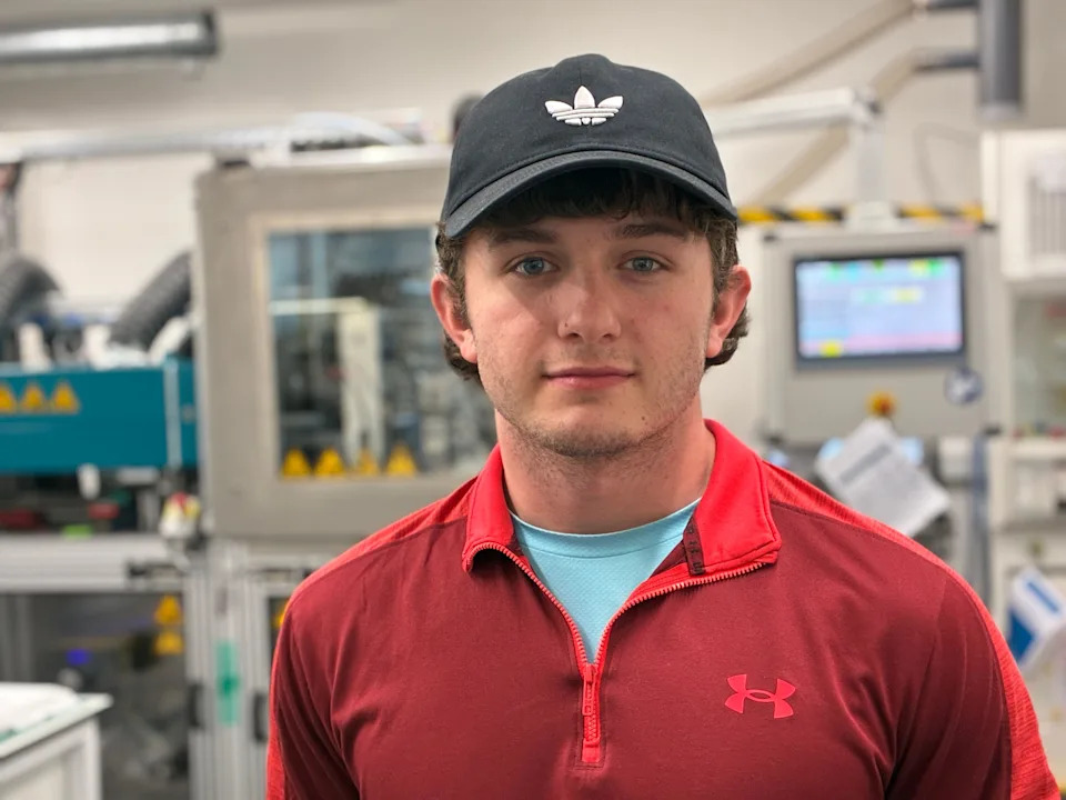 Jack Mason-Young looks at the camera. He has brown hair and is wearing a black baseball cap and red sweater. Behind him is lab machinery.