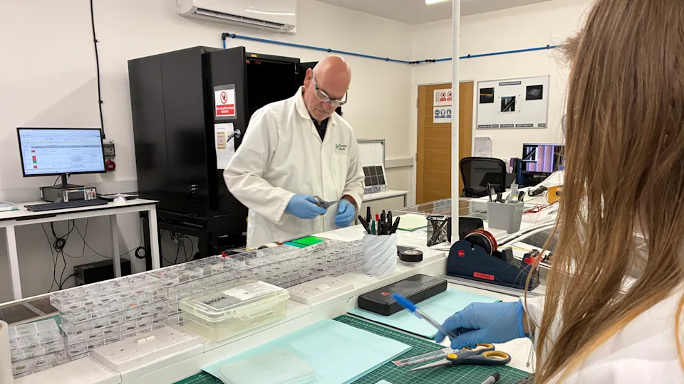 A man in a white lab coat works at a table in a lab setting.
