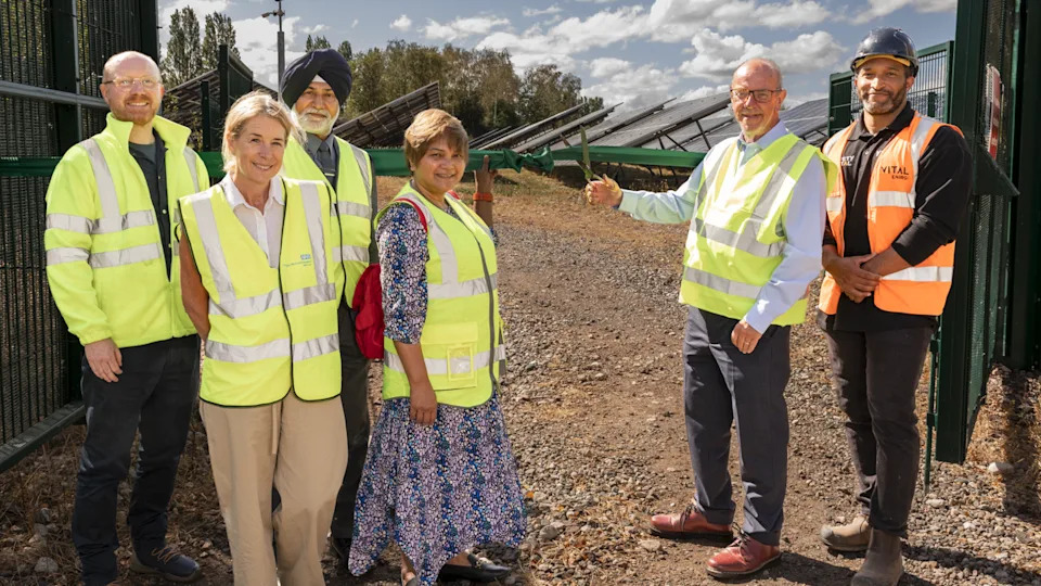 Six people in hi-vis jackets are by a green ribbon and what appears to be an open green gate on the far right. Two men and two women are on the left and one of the two men on the right is holding scissors around the ribbon.